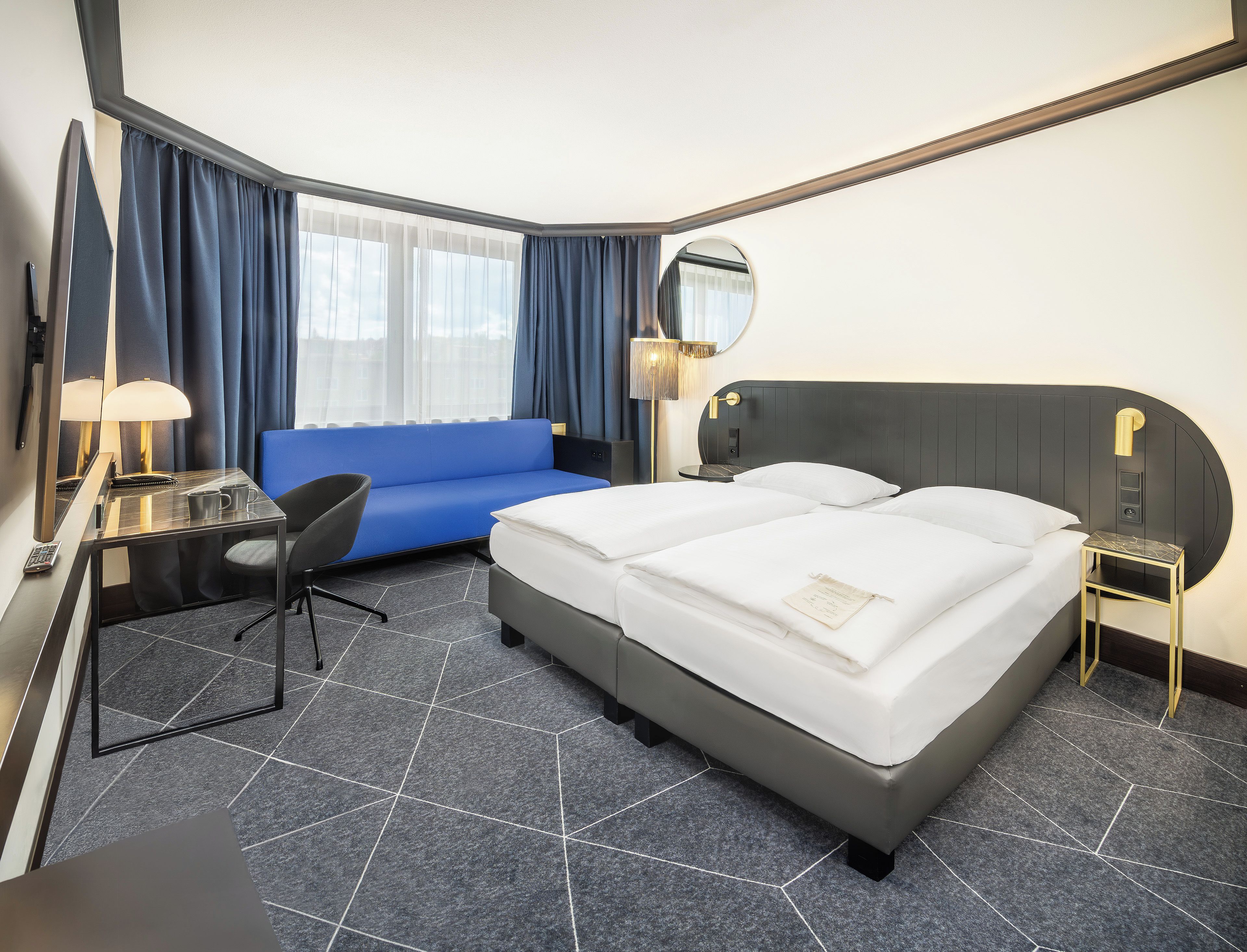 A hotel room with a bed, a desk, a chair, a sofa, a nightstand, a lamp, and a mirror.