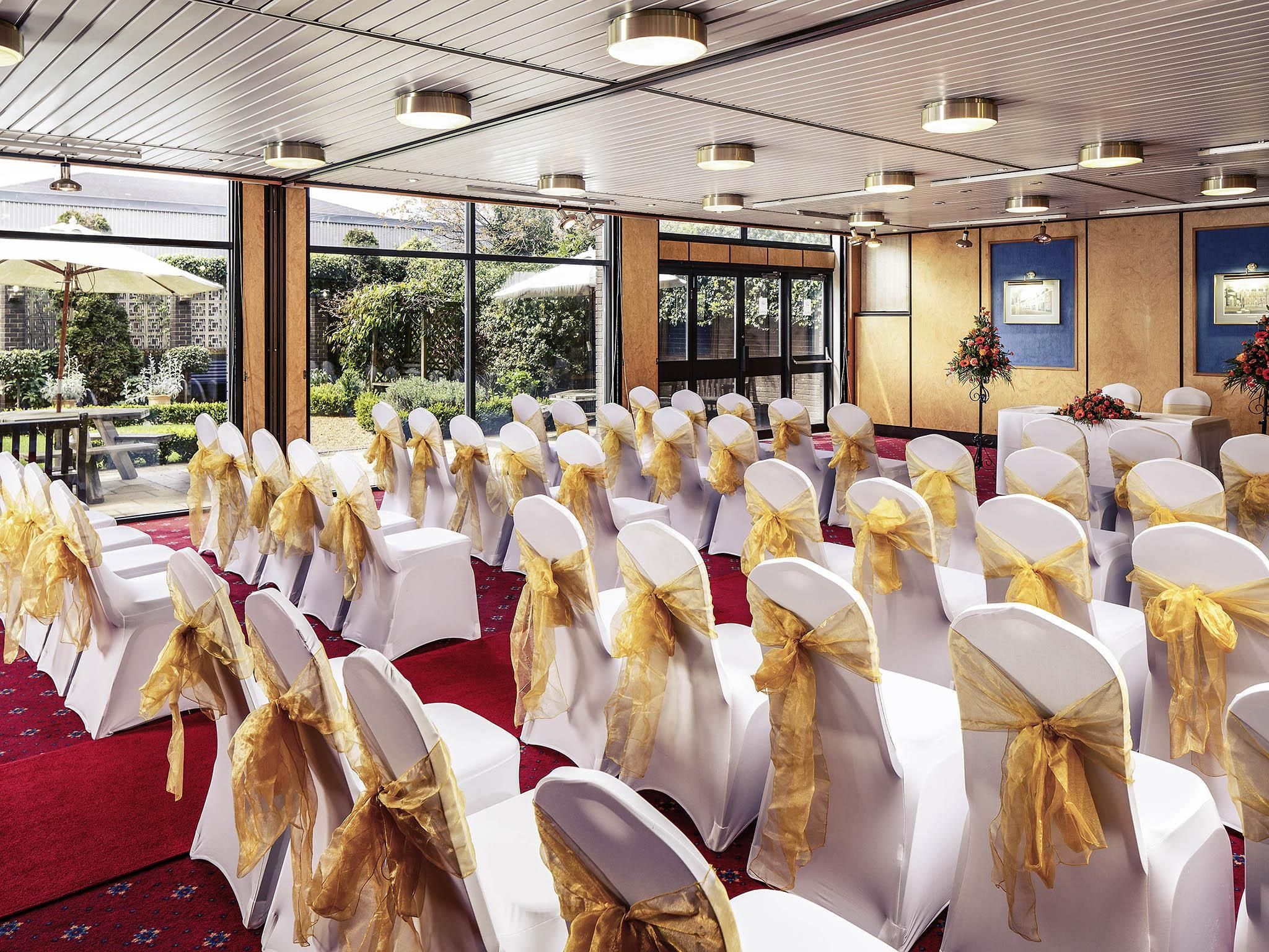 Rows of white chairs with gold sashes, arranged in a banquet hall with large windows and a red carpet.