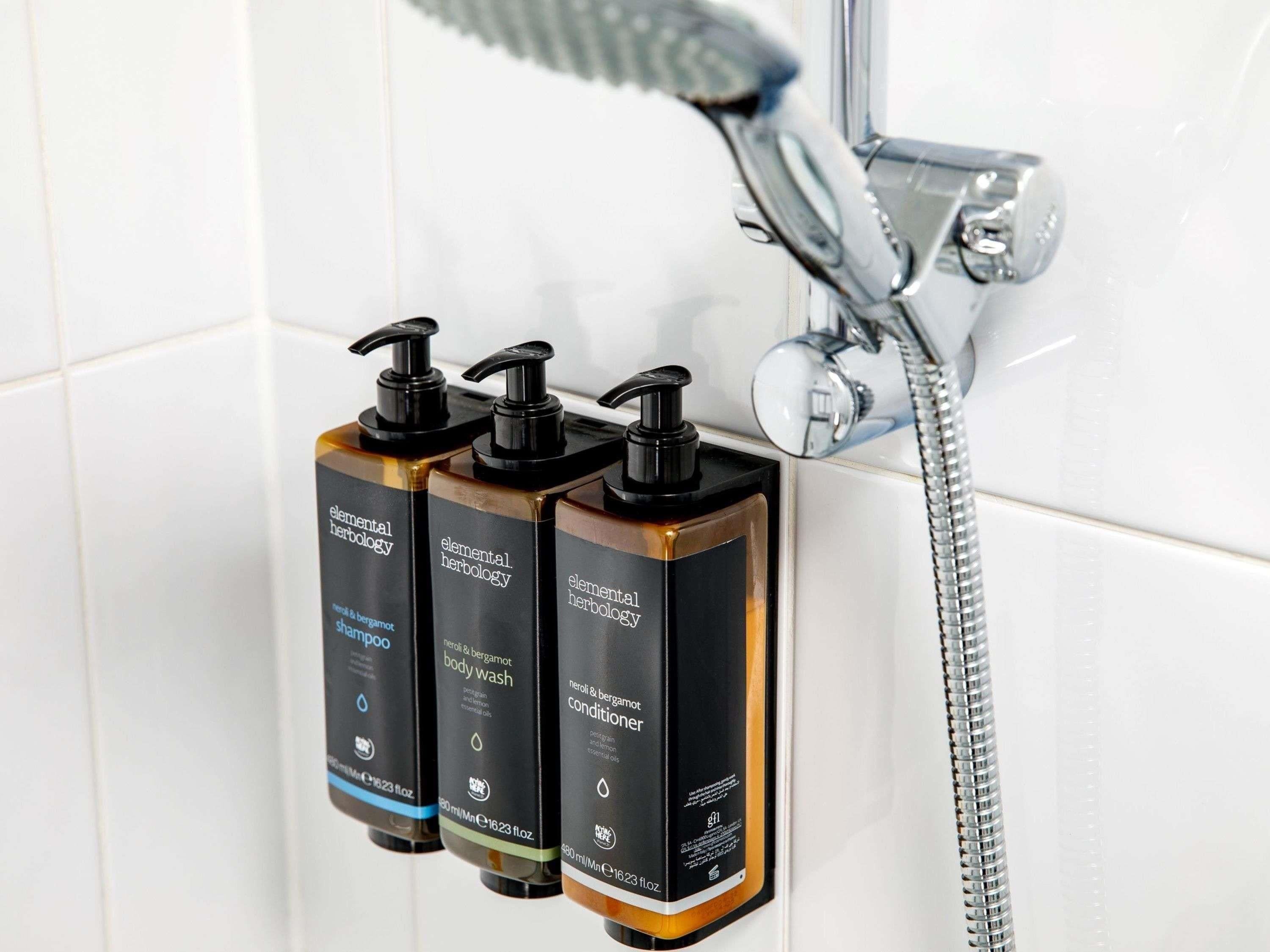 Tres botellas de champú, gel de baño y acondicionador de Elemental Herbology sobre una pared de azulejos cerca de una ducha.