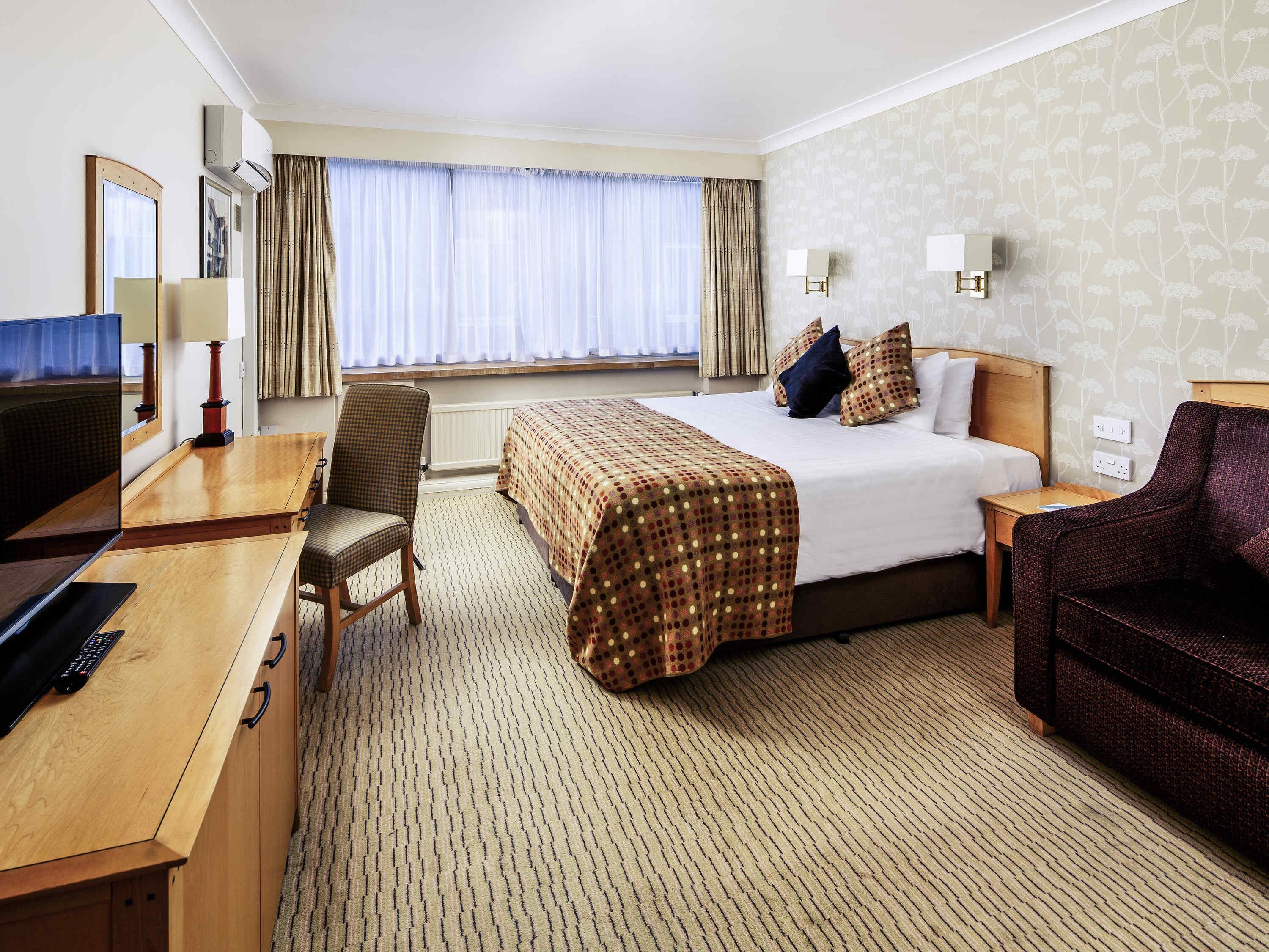 A hotel room with a bed, a desk, a chair, a TV, and a window with curtains.