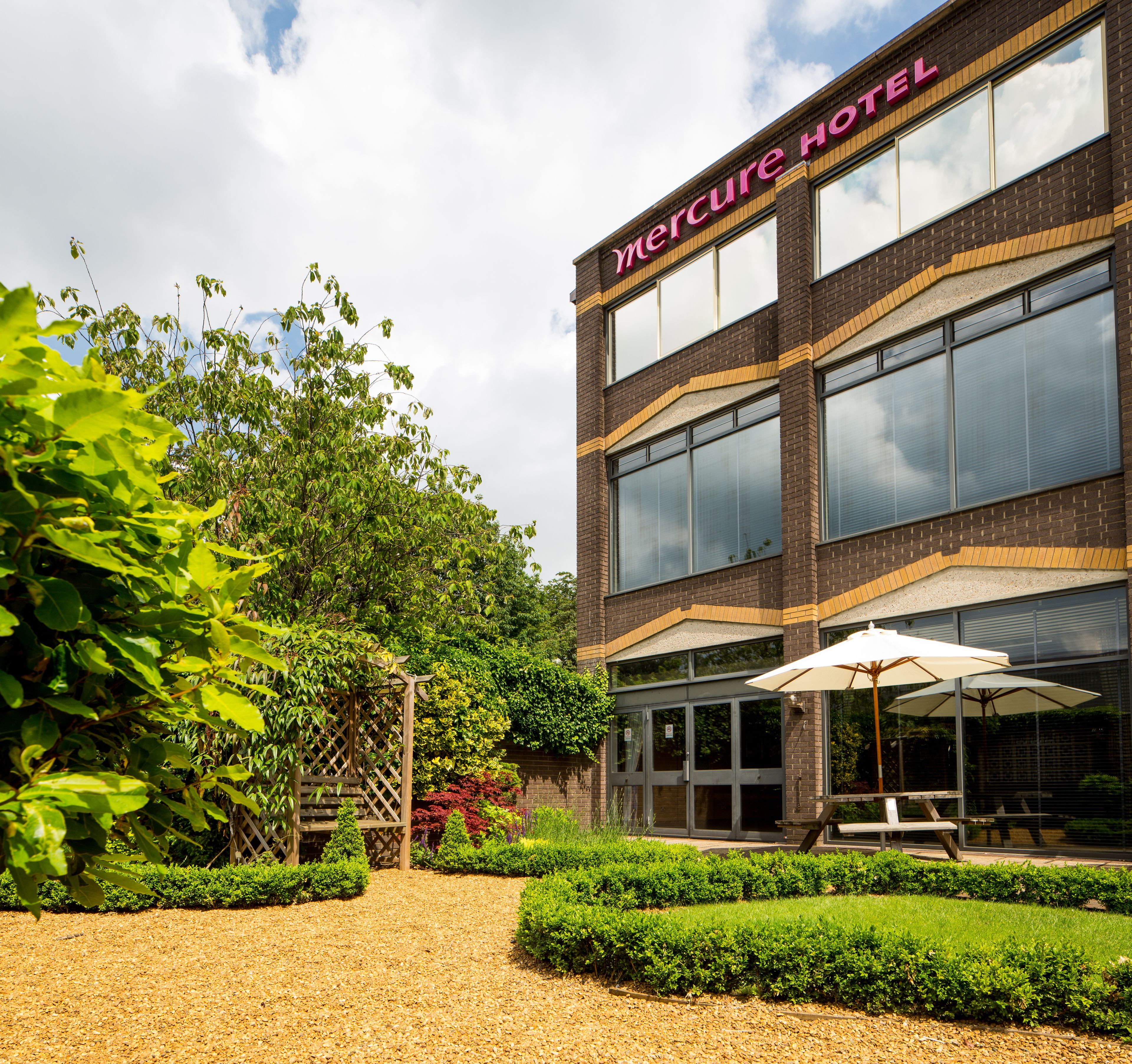 A Mercure Hotel with a modern brick facade, large windows, and a well-maintained garden with a gravel path.