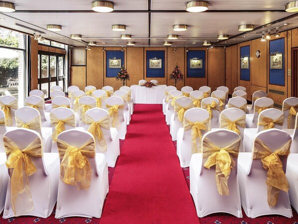 Un salón de conferencias con filas de sillas blancas con cintas amarillas, una alfombra roja y cuadros enmarcados en las paredes.