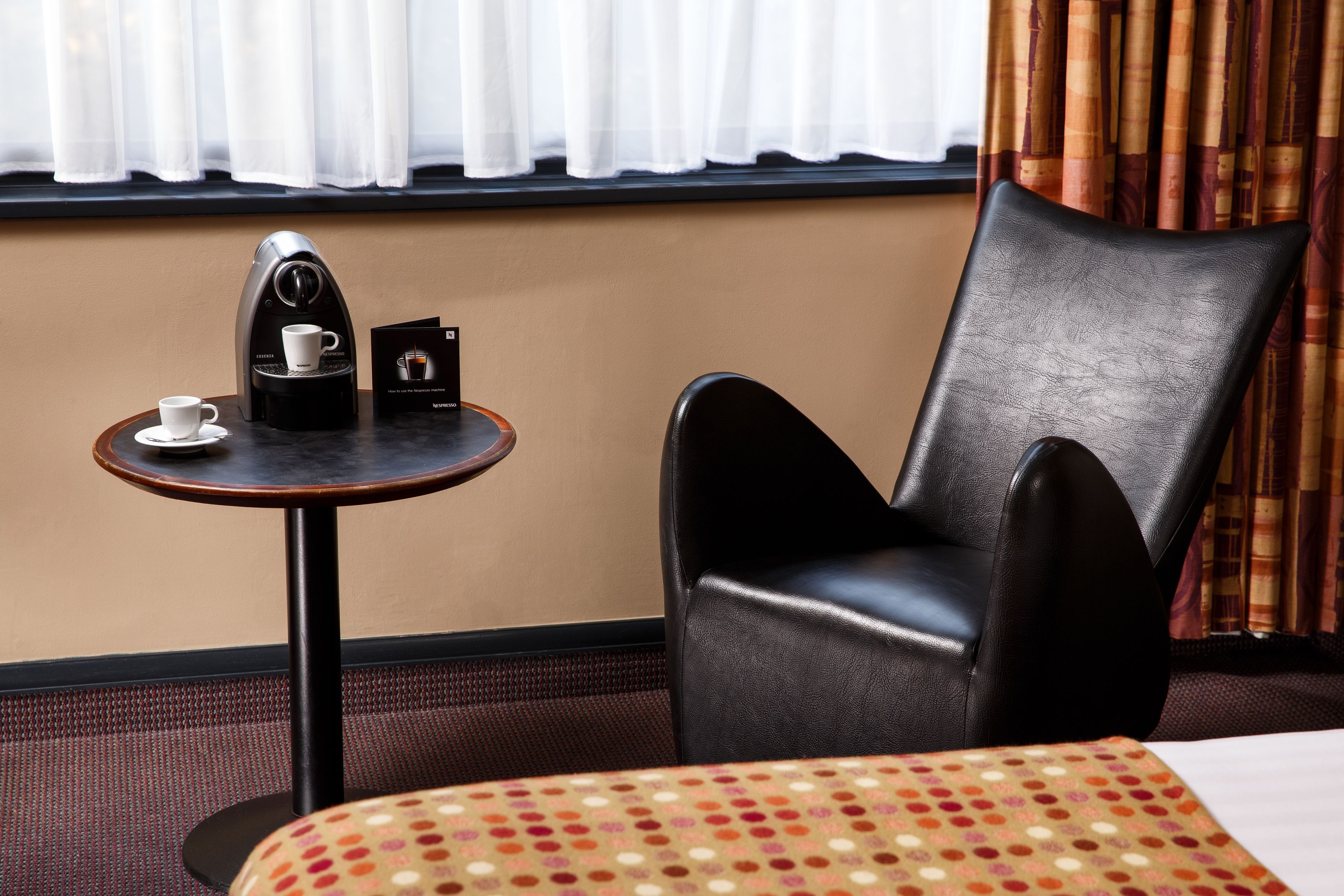 A hotel room with a single black leather chair, a small round table with a coffee maker and a cup, and a bed with a patterned bedspread.