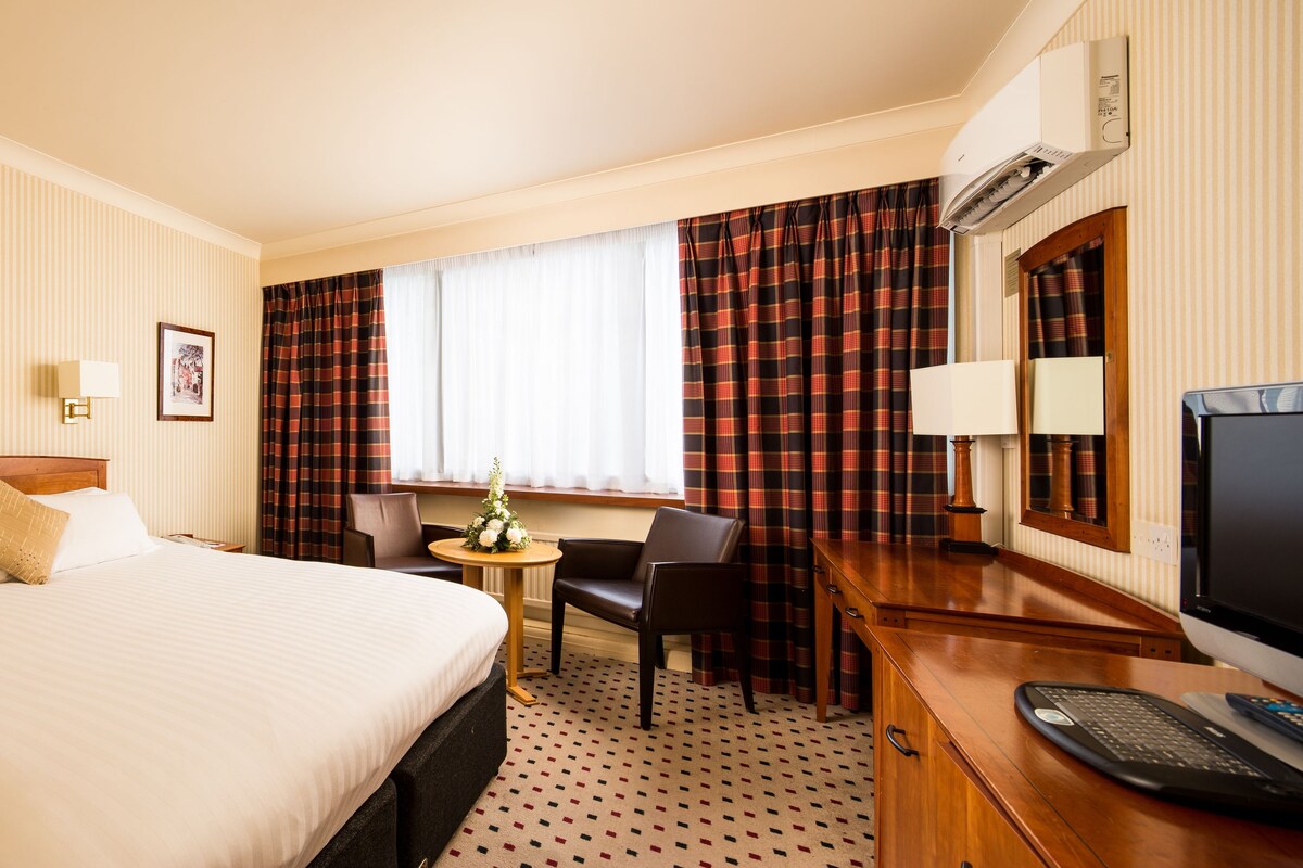 A hotel room with a large bed, a desk with a computer, a chair, a small table with flowers, and a television.