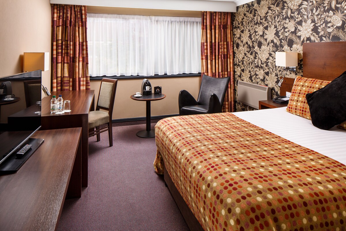 A hotel room with a bed, a desk, a chair, a small table, and a window with curtains.