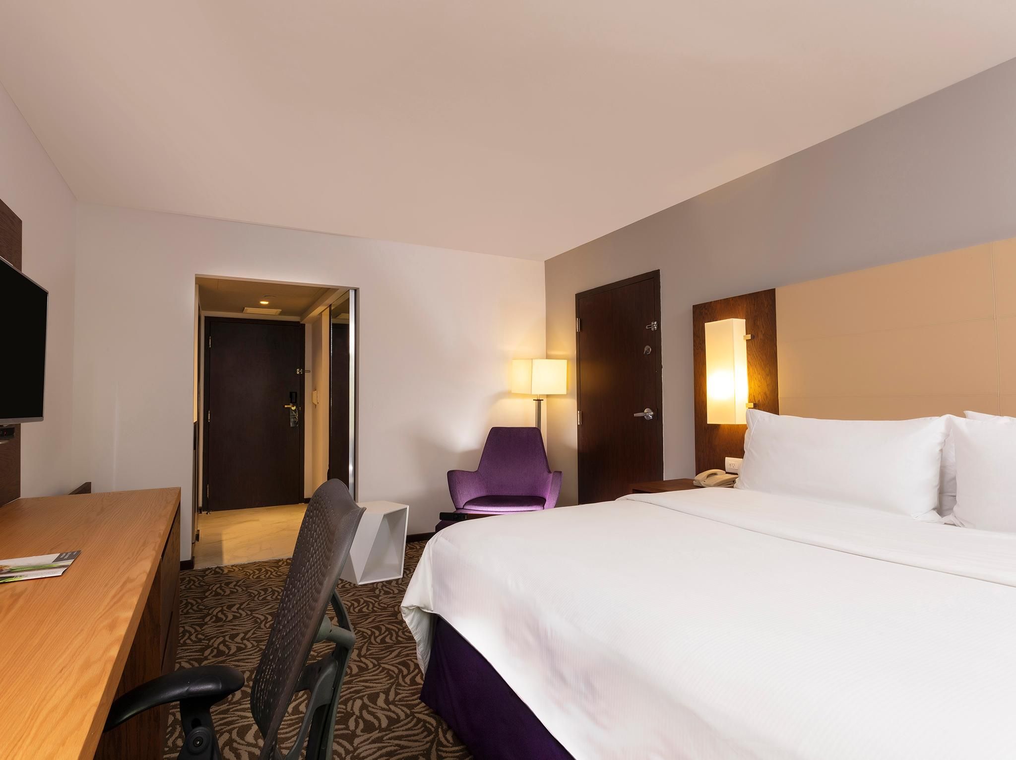 A hotel room with a bed, a desk, a chair, a lamp, and a door.