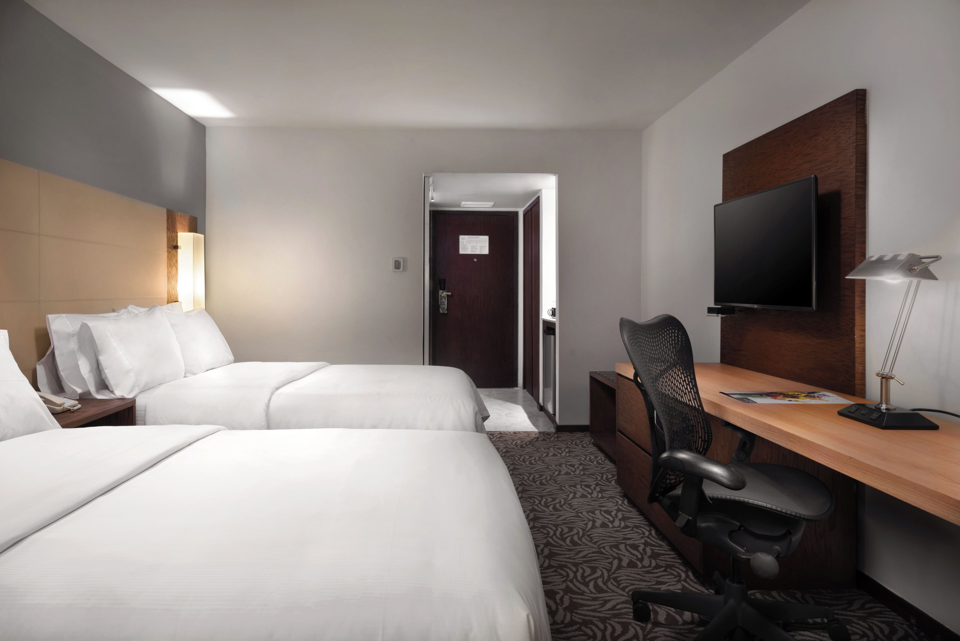 A hotel room with two beds, a desk, a chair, a television, and a door.
