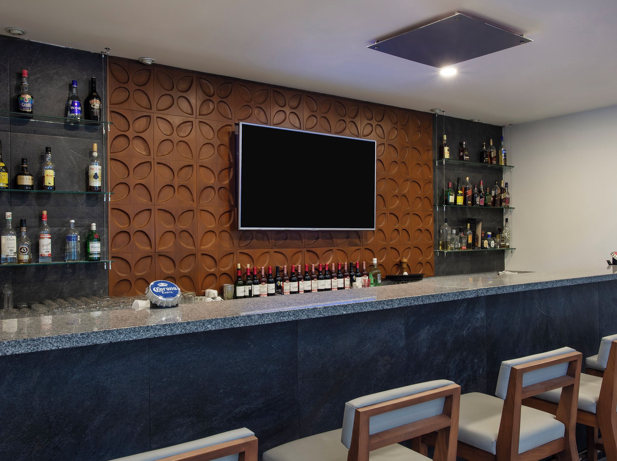 Un espace bar avec un téléviseur à écran plat fixé sur un mur recouvert de panneaux en bois, un comptoir orné de différentes bouteilles et des tabourets de bar.