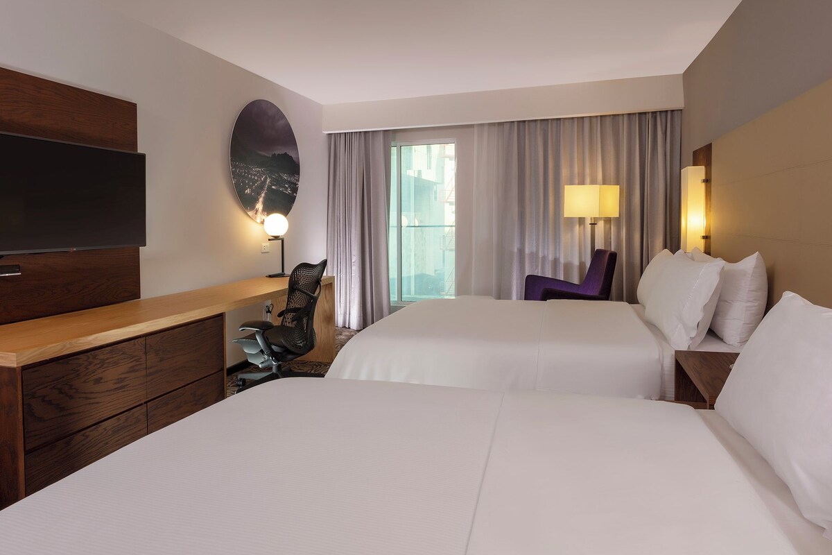 A hotel room with two beds, a desk, a chair, a TV, and a window with curtains.