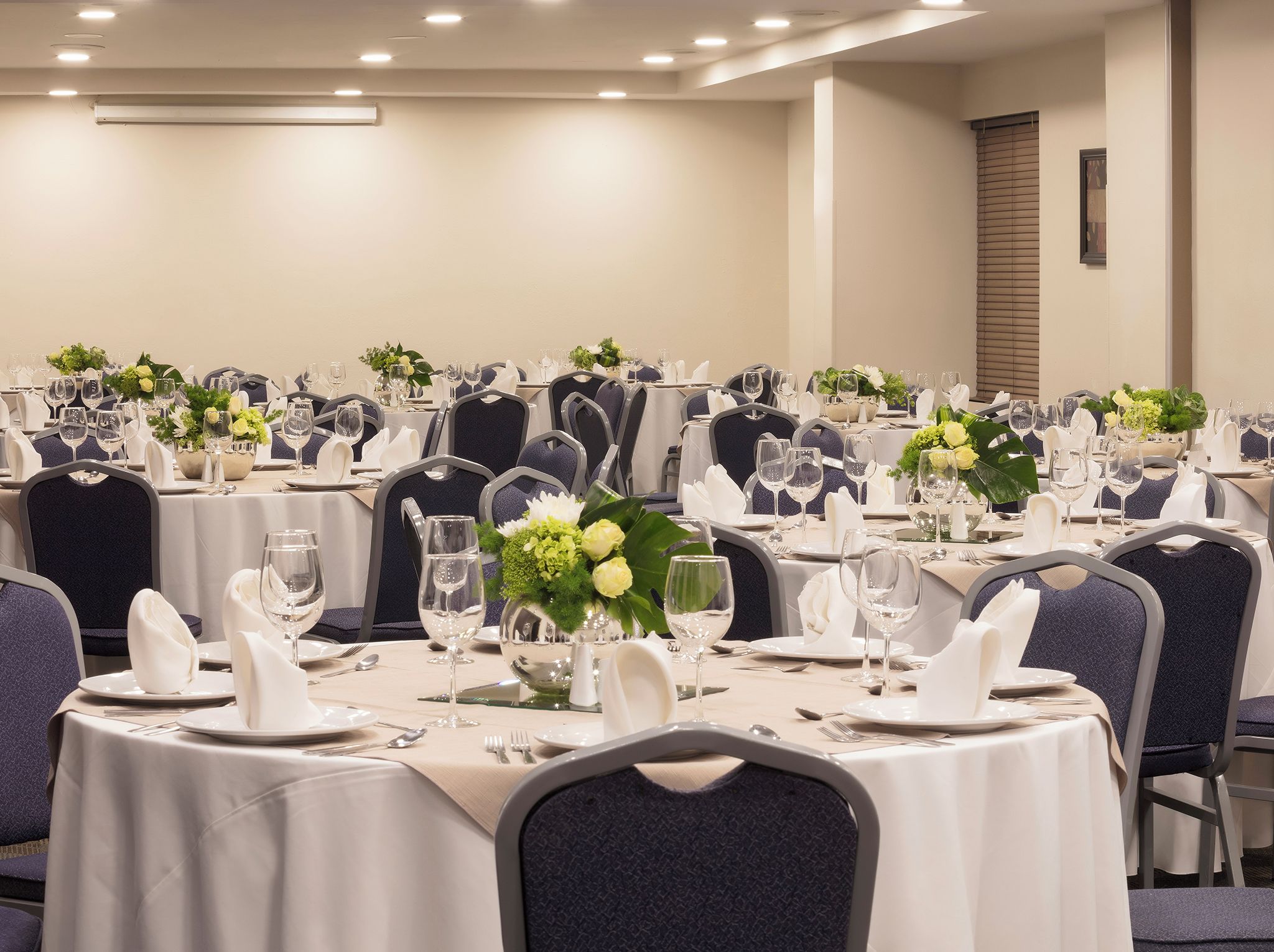 Un salón de conferencias con varias mesas dispuestas para comer, cada una con mantel blanco, sillas y centros de mesa con flores.