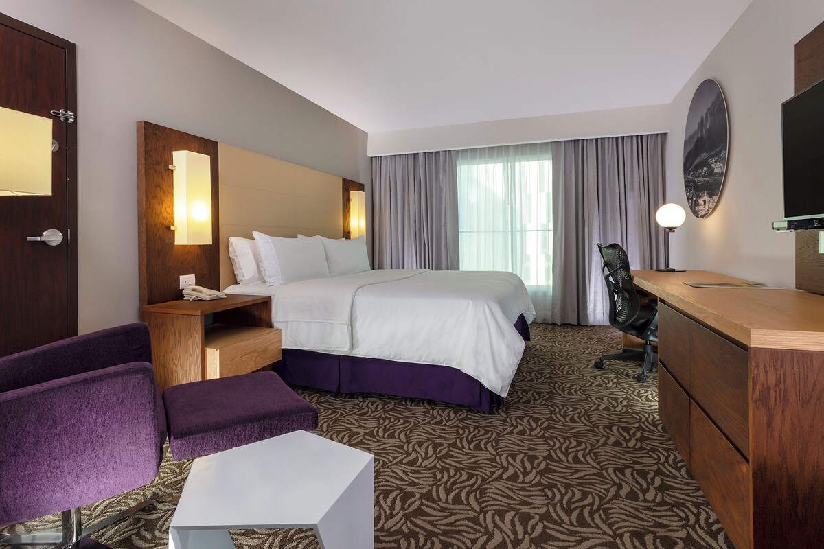 A hotel room with a bed, a desk, a chair, a small table, a mirror, and a television.