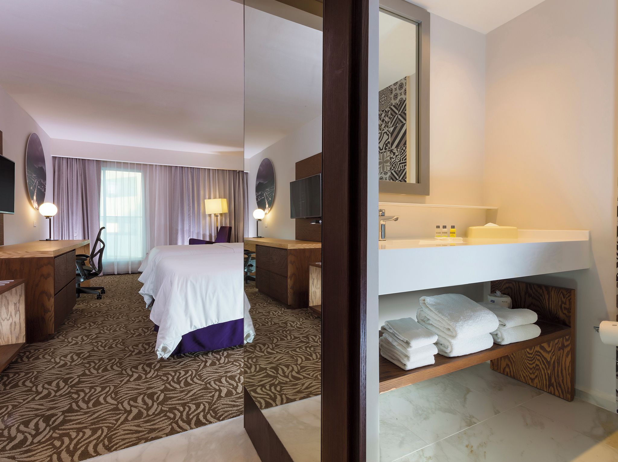 Habitación de hotel con cama, escritorio, silla, televisor y baño con lavamanos y toallas.