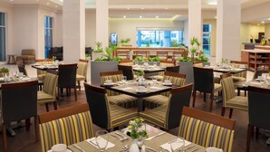 Un vaste espace dédié à la salle à manger, avec de nombreuses tables et chaises, une colonne centrale et une vue sur l’extérieur grâce à de grandes fenêtres.