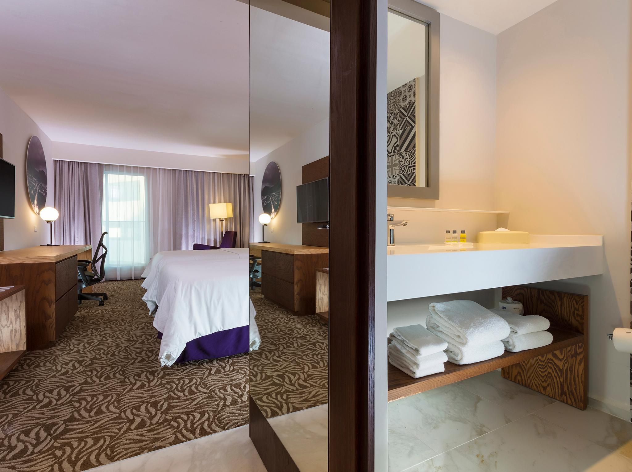A hotel room with a bed, desk, chair, lamp, TV, and a bathroom with a sink and towels.