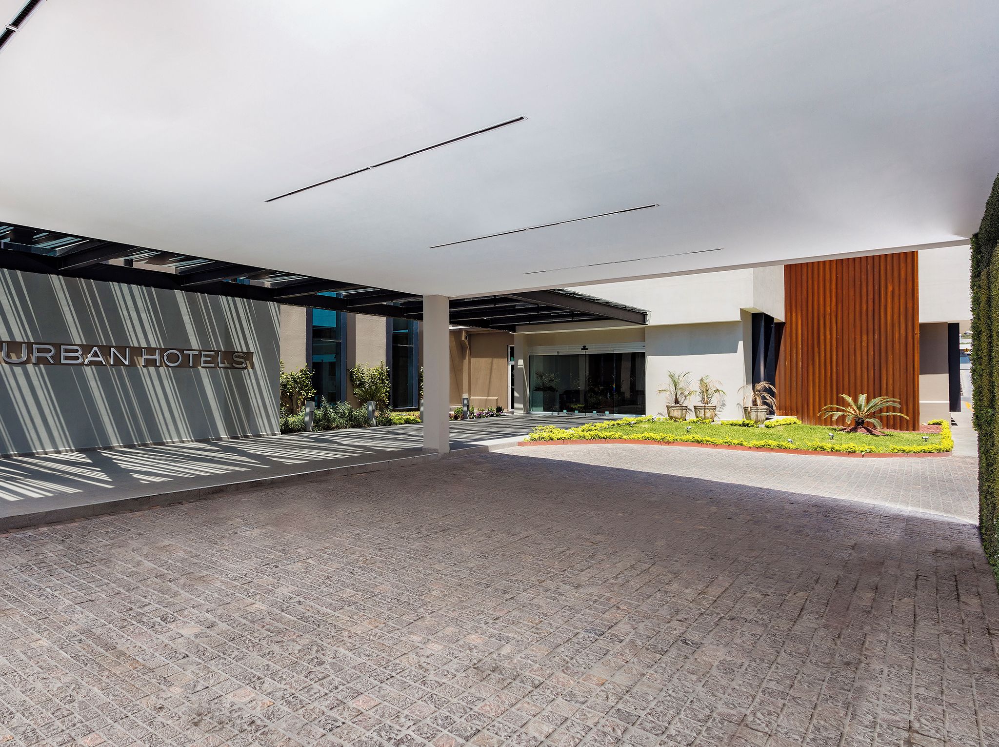 A modern hotel entrance with a covered walkway, a sign reading 'URBAN HOTELS', and a landscaped garden area.