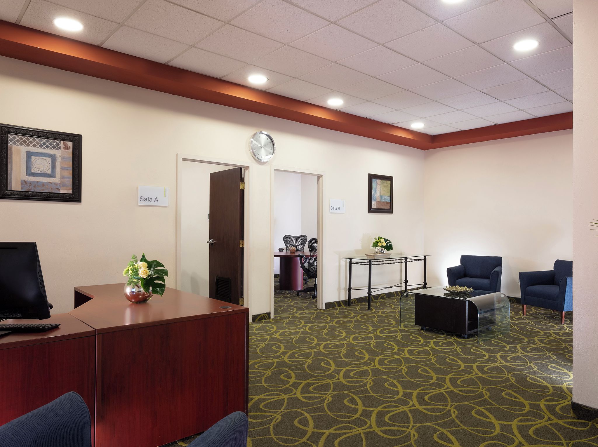 A conference room with a wooden desk, two blue chairs, a glass coffee table, and a clock on the wall.