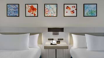 A hotel room with two beds, a bedside table with a phone and a small clock, and four framed artworks on the wall.