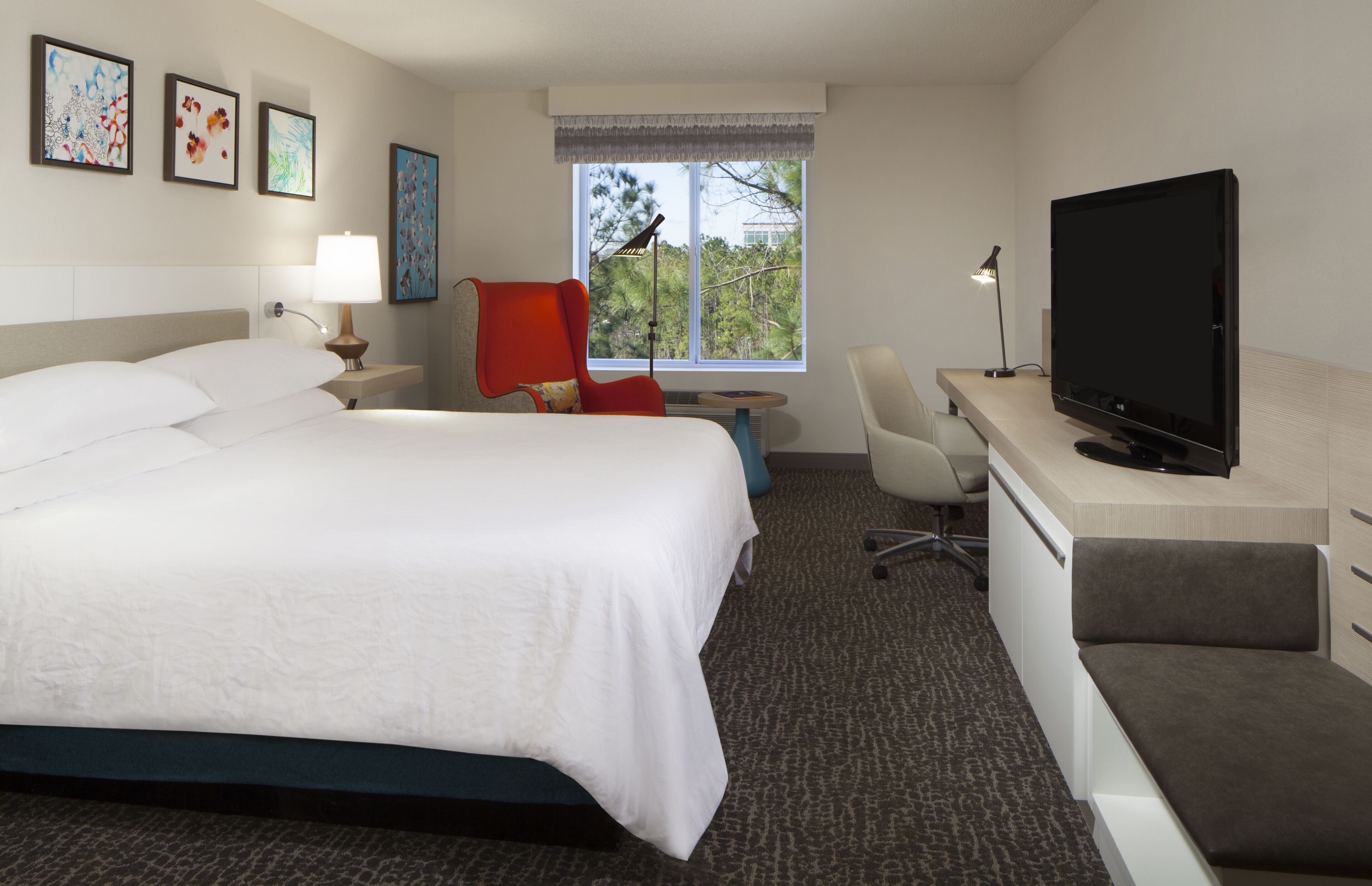 A hotel room with a bed, desk, TV, chair, and artwork on the wall.
