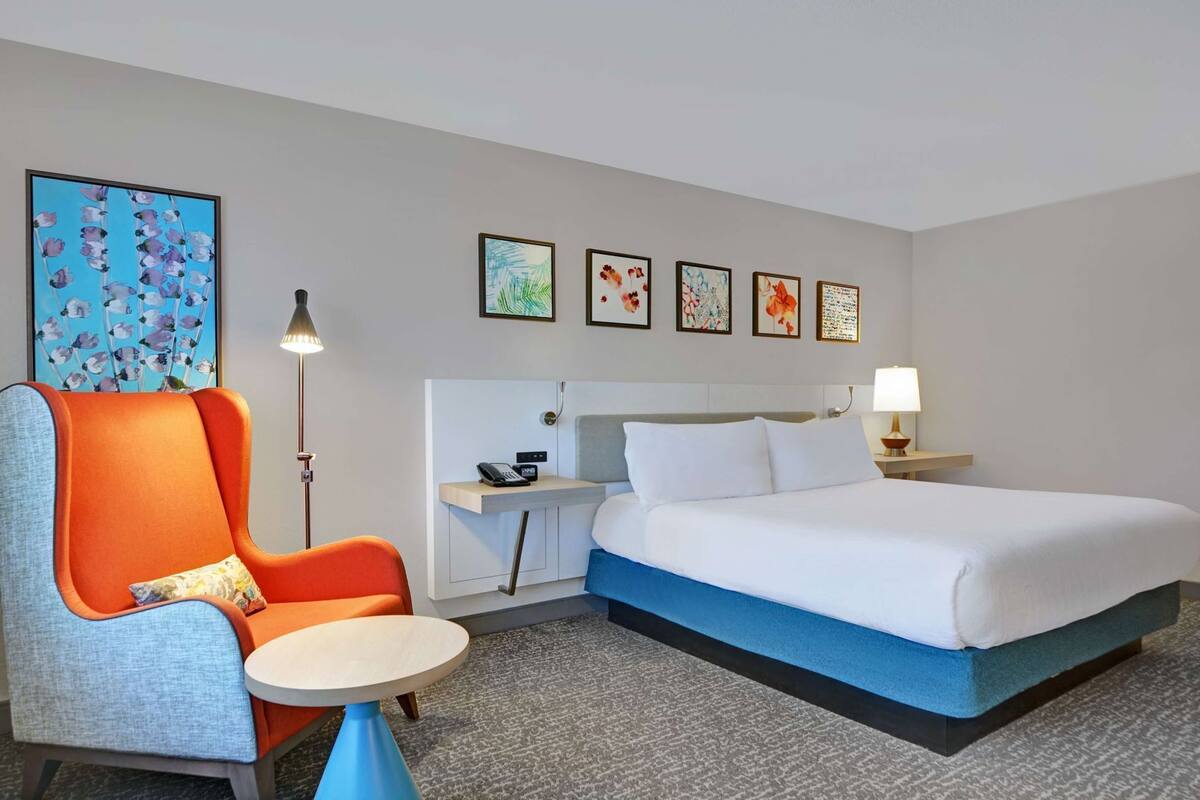 A hotel room with a bed, a desk with a phone, a chair, a small table, and artwork on the wall.