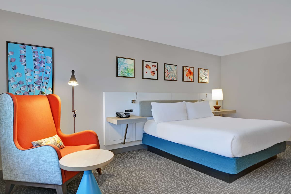 Une chambre d’hôtel avec un lit, un fauteuil orange, une petite table ronde, un bureau avec une lampe et quatre tableaux encadrés accrochés au mur.