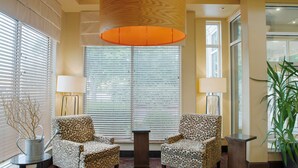 A lobby with two patterned armchairs, a round table, a large window with blinds, a hanging orange lamp, and a potted plant. - Hilton Garden Inn Bentonville Rogers (Bentonville)