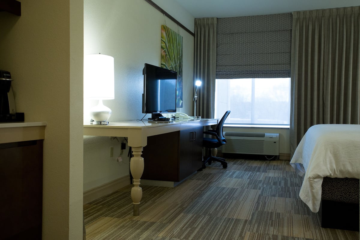 Une chambre d’hôtel comprenant un lit, un bureau avec un ordinateur et une lampe, une chaise, une fenêtre avec des rideaux et un appareil de climatisation.