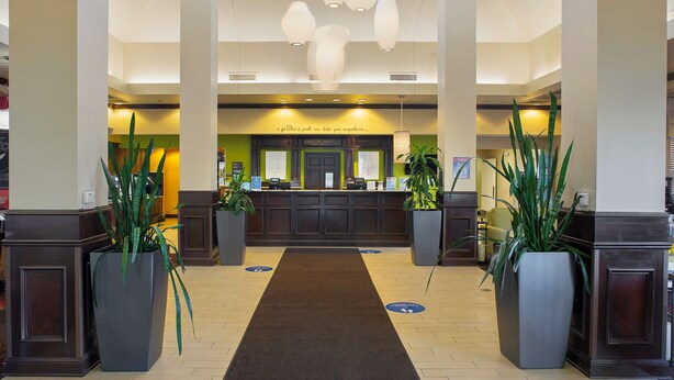 A hotel lobby with a reception desk, large potted plants, and a black carpet leading to the desk. - Hilton Garden Inn Bentonville Rogers (Bentonville)
