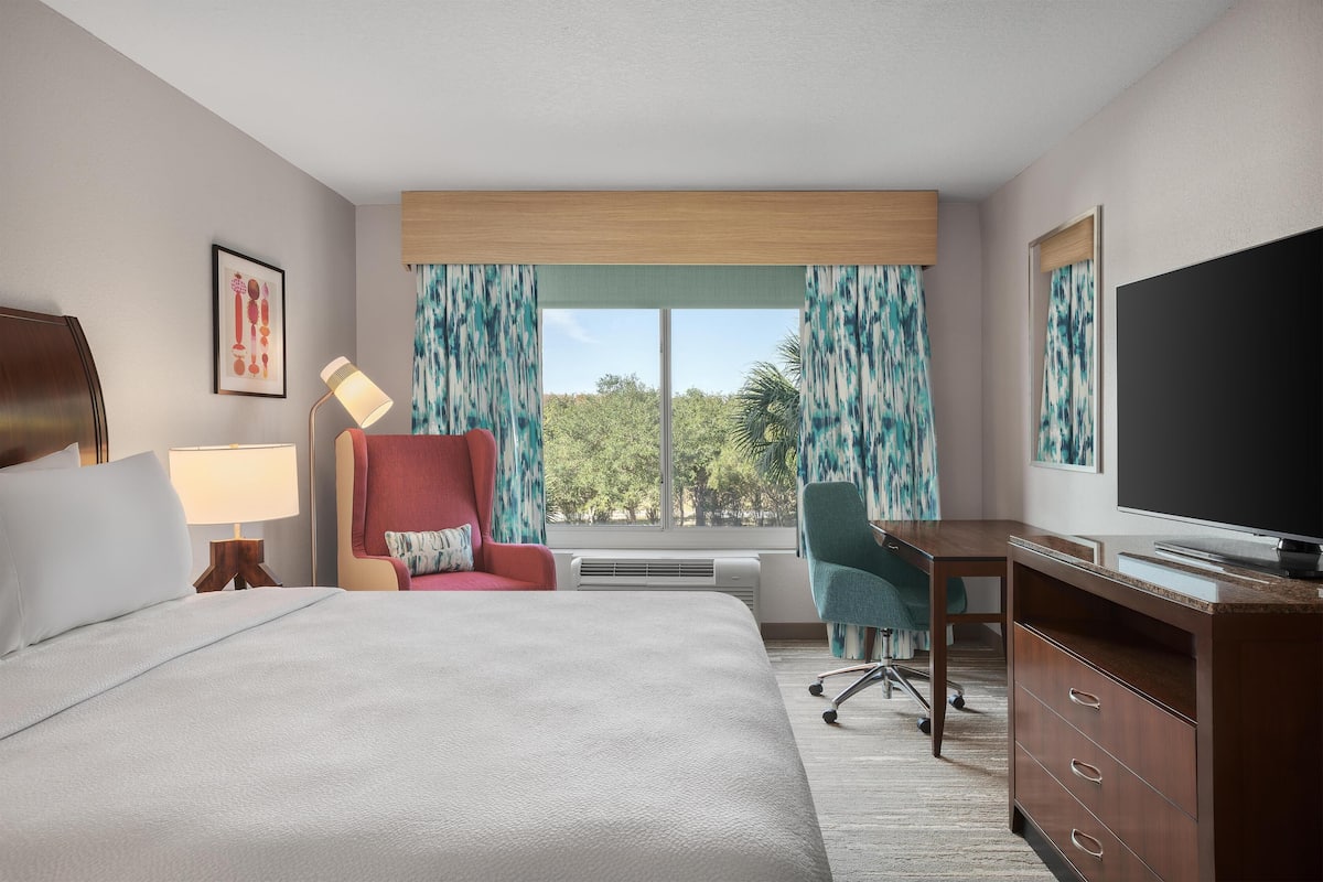 Habitación de hotel con una cama grande, un escritorio, una silla, un televisor y una ventana con cortinas.