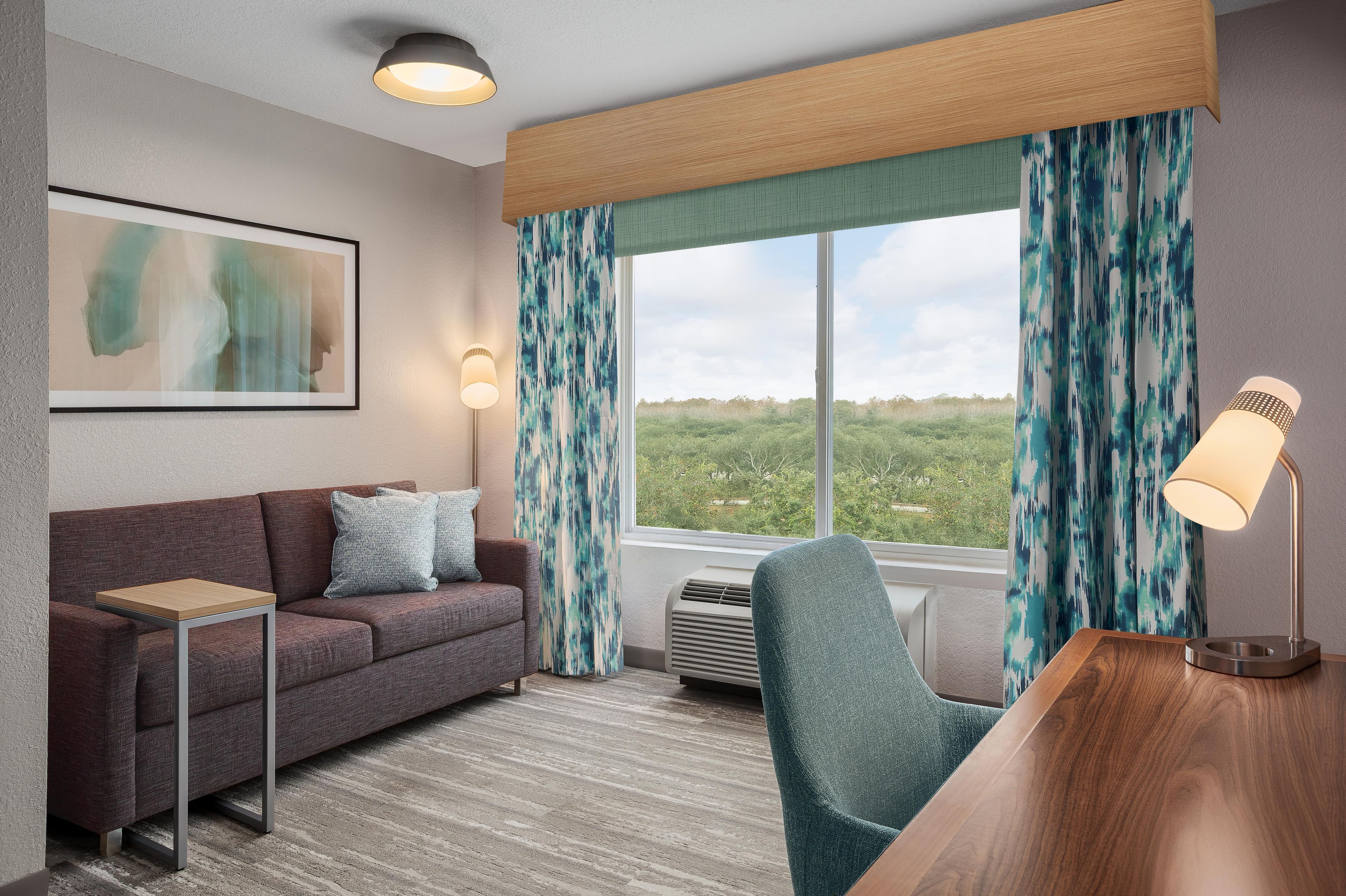 Een hotelkamer met een bank, een stoel, een bureau, een lijst met kunstwerk en een groot raam met gordijnen.