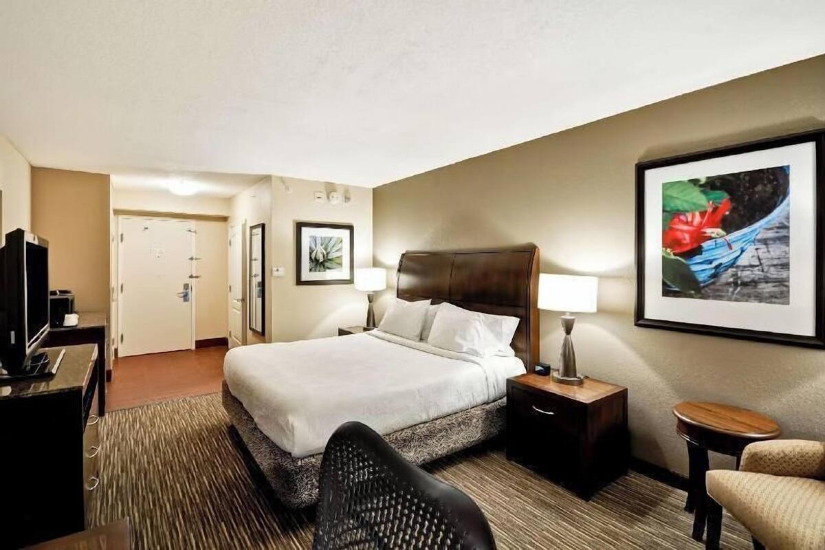 Habitación de hotel con cama, escritorio con computadora, silla, mesita pequeña y un cuadro en la pared.