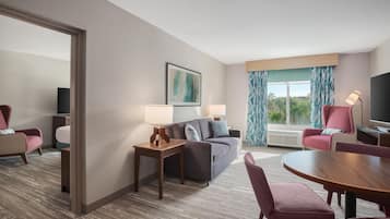A hotel room with a sofa, two armchairs, a dining table, a TV, and a window with curtains.