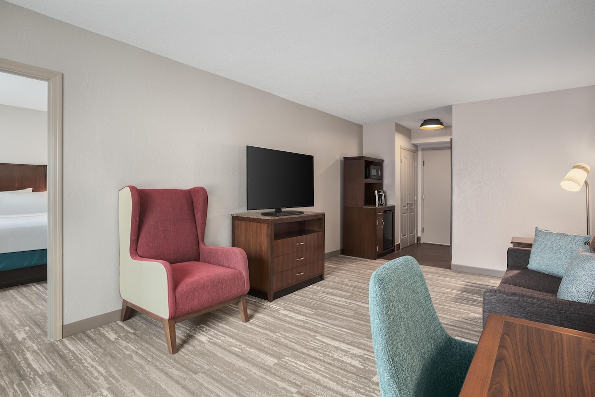 Habitación de hotel con un sillón rojo, un televisor sobre un mueble de madera, una mesita con una lámpara y vistas al pasillo.