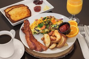 Un plato de desayuno con tostadas, tocino, papas, huevos revueltos y un vaso de jugo de naranja.