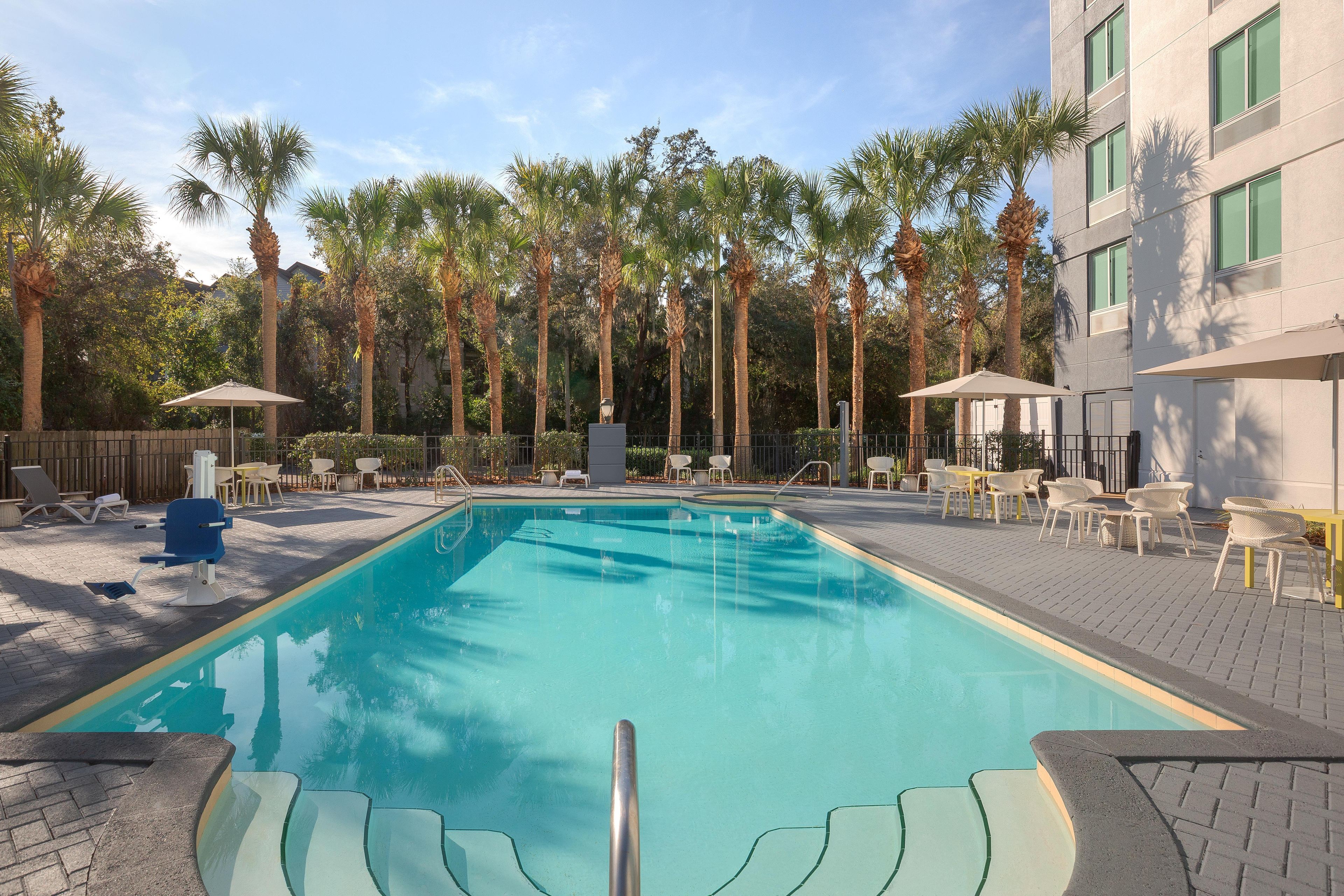 A swimming pool with lounge chairs and umbrellas, surrounded by palm trees and a building.