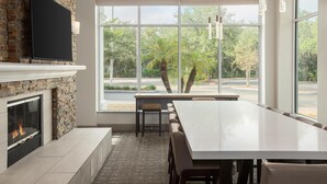 Un comedor moderno con una gran mesa, una chimenea y ventanas amplias que dan vistas a árboles y una carretera.