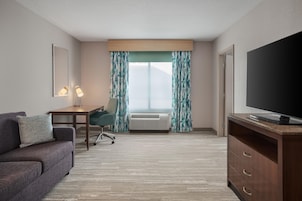 A hotel room with a sofa, a desk with a lamp, a television, and a large window with curtains.