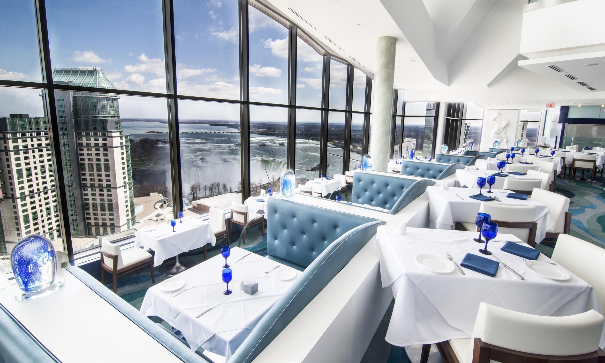 A high-rise restaurant with large windows offering a city view, multiple dining tables, and blue cushioned seating.