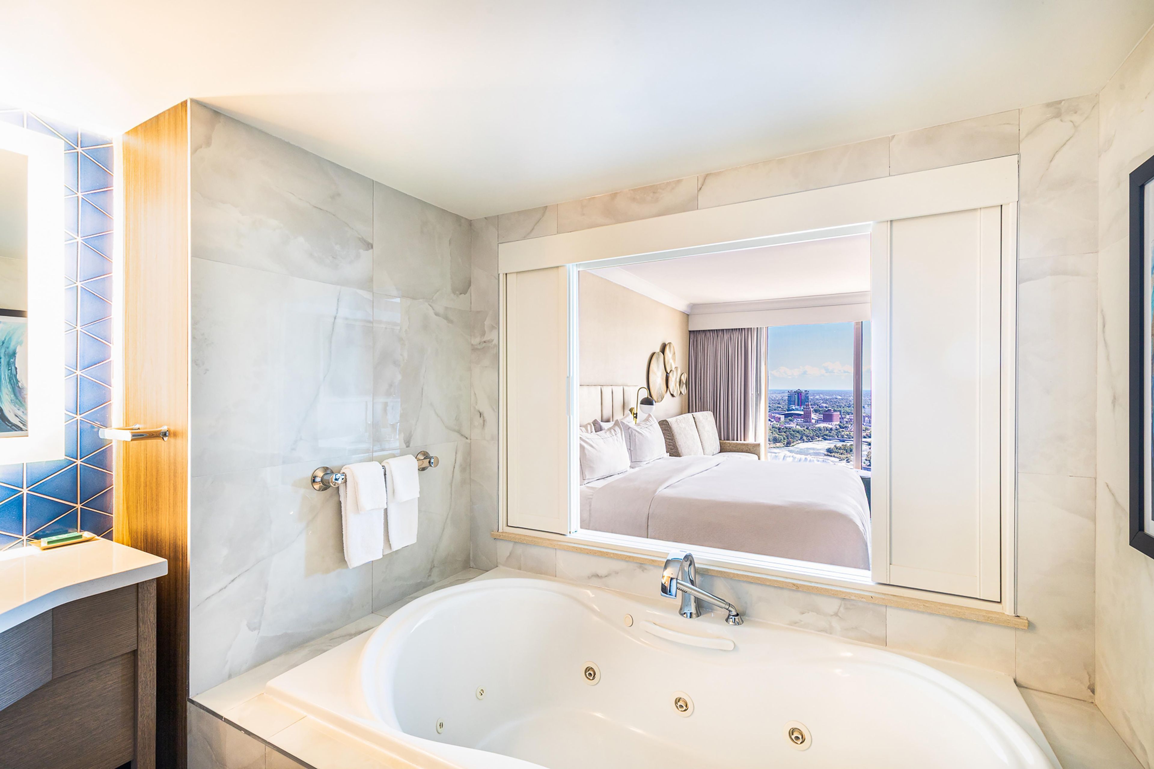 Une salle de bain moderne avec une grande baignoire, un miroir qui reflète une chambre à coucher et une vue sur la ville.