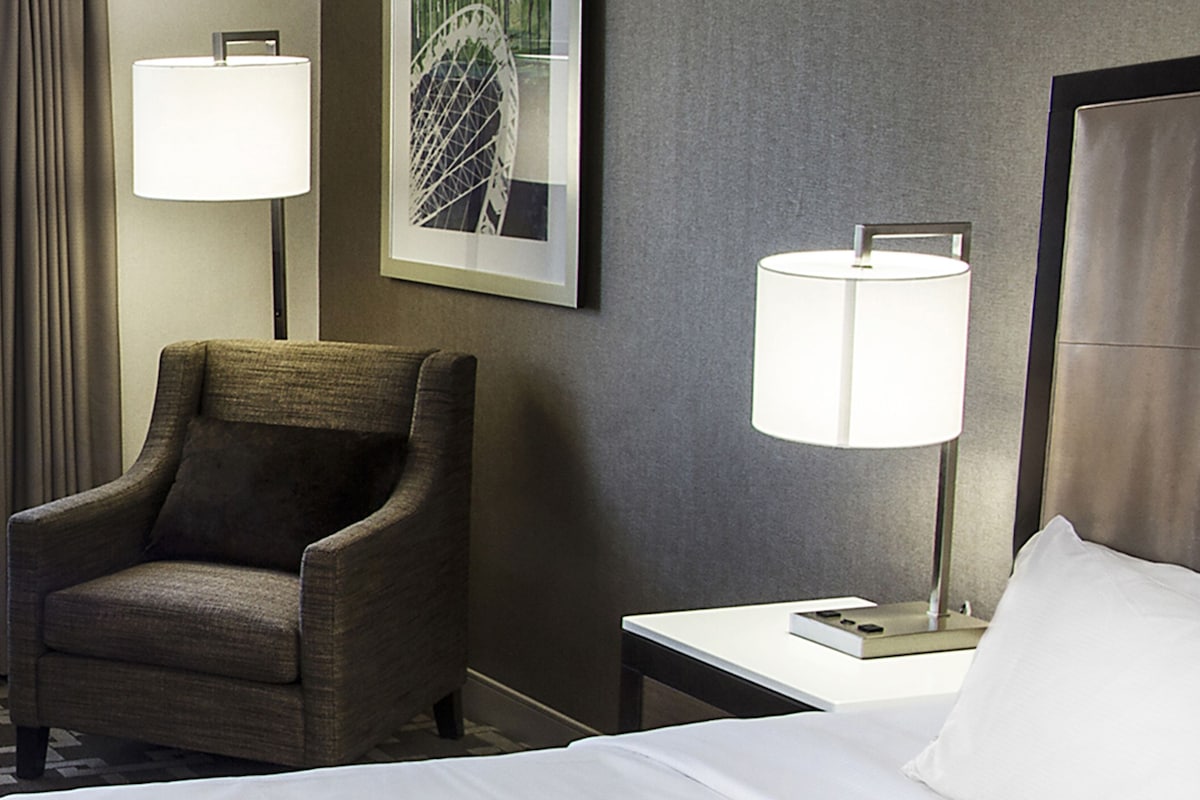 A hotel room with a bed, a chair, a nightstand, a lamp, and a framed picture on the wall.