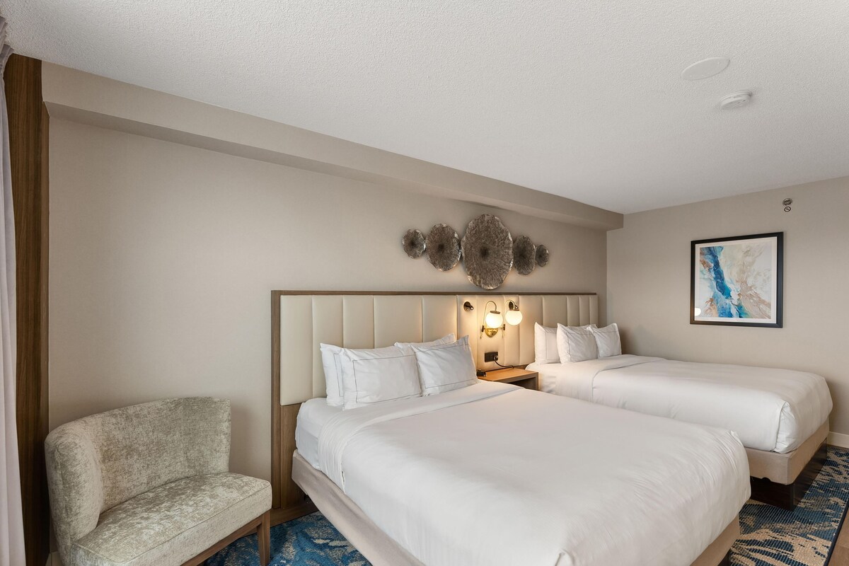 A hotel room with two beds, a chair, a wall-mounted light fixture, and a framed picture.
