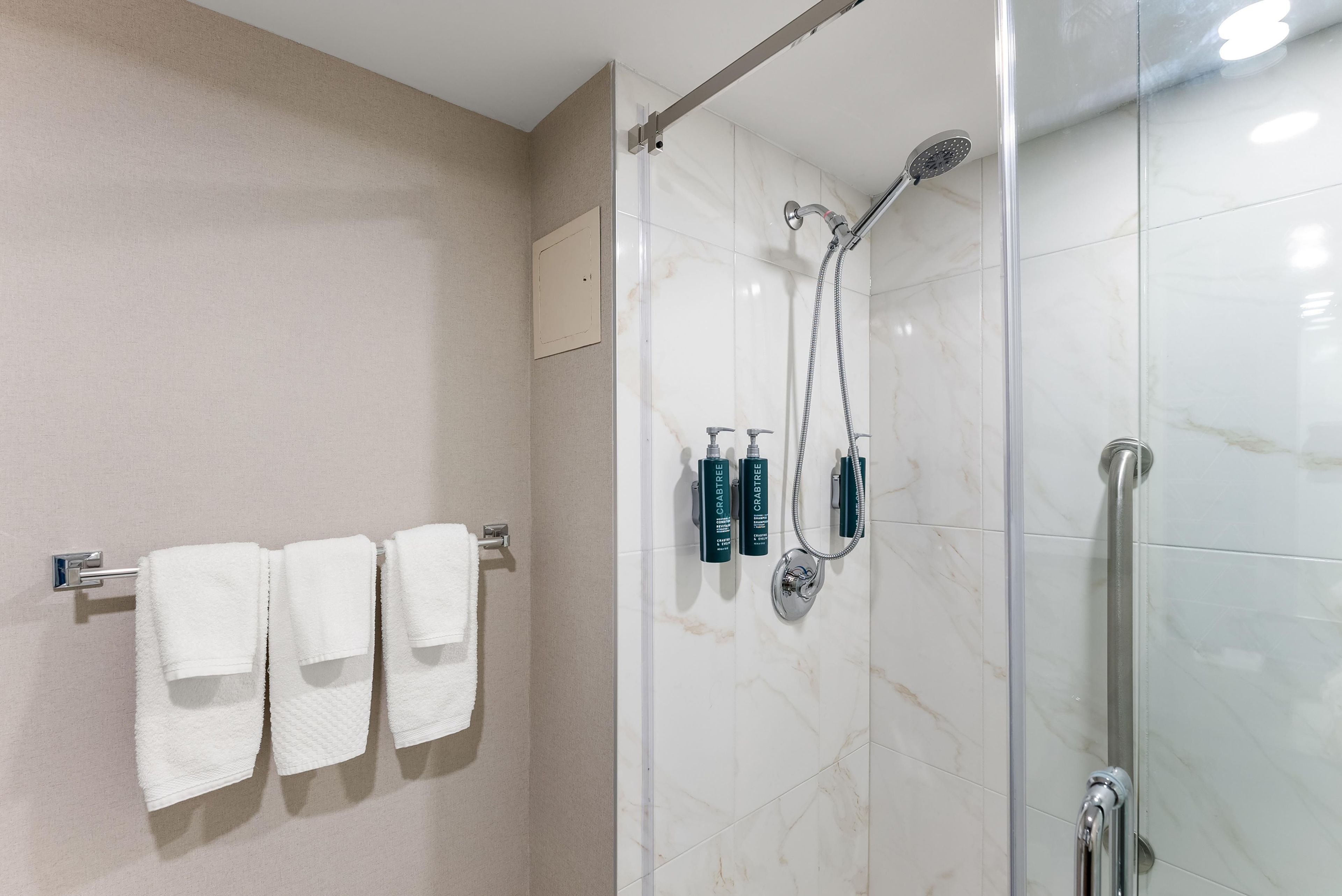 Une douche équipée d’un pommeau de douche à main et d’un robinet mural, d’une porte en verre et de trois serviettes blanches suspendues à un porte-serviettes.
