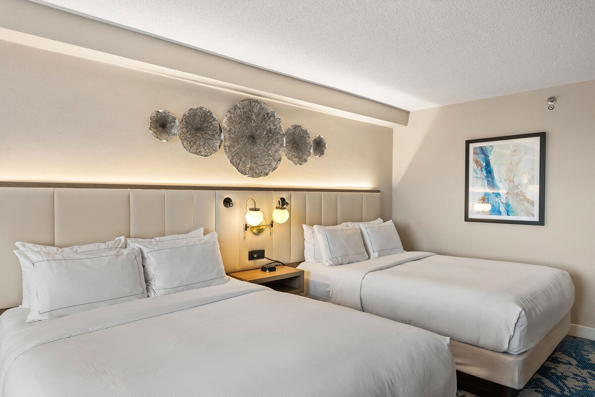 A hotel room with two beds, a nightstand, wall sconces, and a framed abstract painting.
