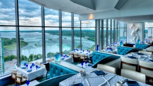 A restaurant with large windows offering a view of a waterfall and landscape. Tables are set with blue napkins and white tablecloths. - Hilton Niagara Falls/Fallsview Hotel & Suites (Niagara Falls)