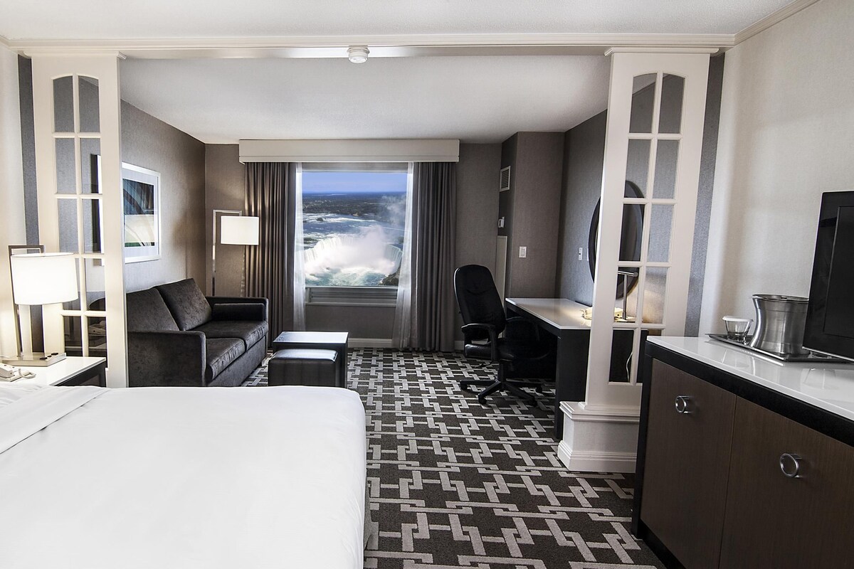 A hotel room with a bed, a desk, a chair, a sofa, a TV, a lamp, and a view of a waterfall.