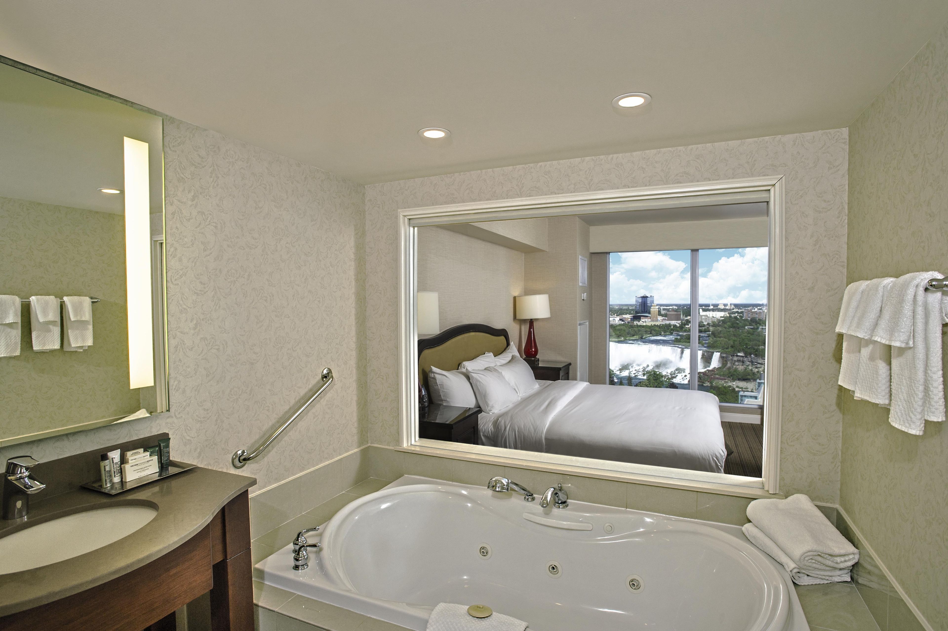 Un baño de hotel con un espejo grande, una bañera, un lavamanos y vistas a la ciudad a través de una ventana.