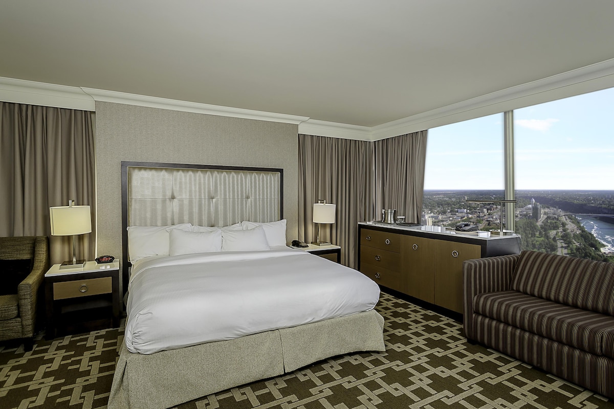 A hotel room with a large bed, a desk, a chair, a dresser, and a view of a cityscape.
