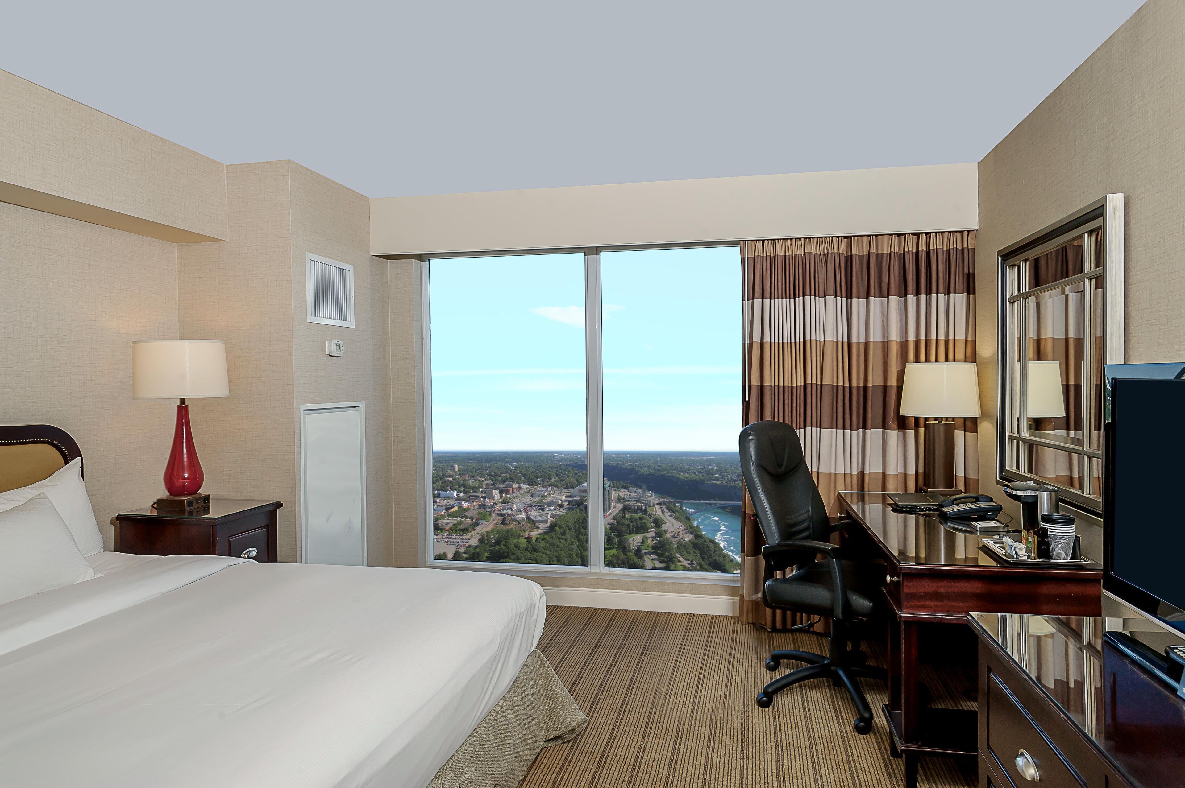 A hotel room with a large bed, a desk with a chair, a TV, and a view of a cityscape.