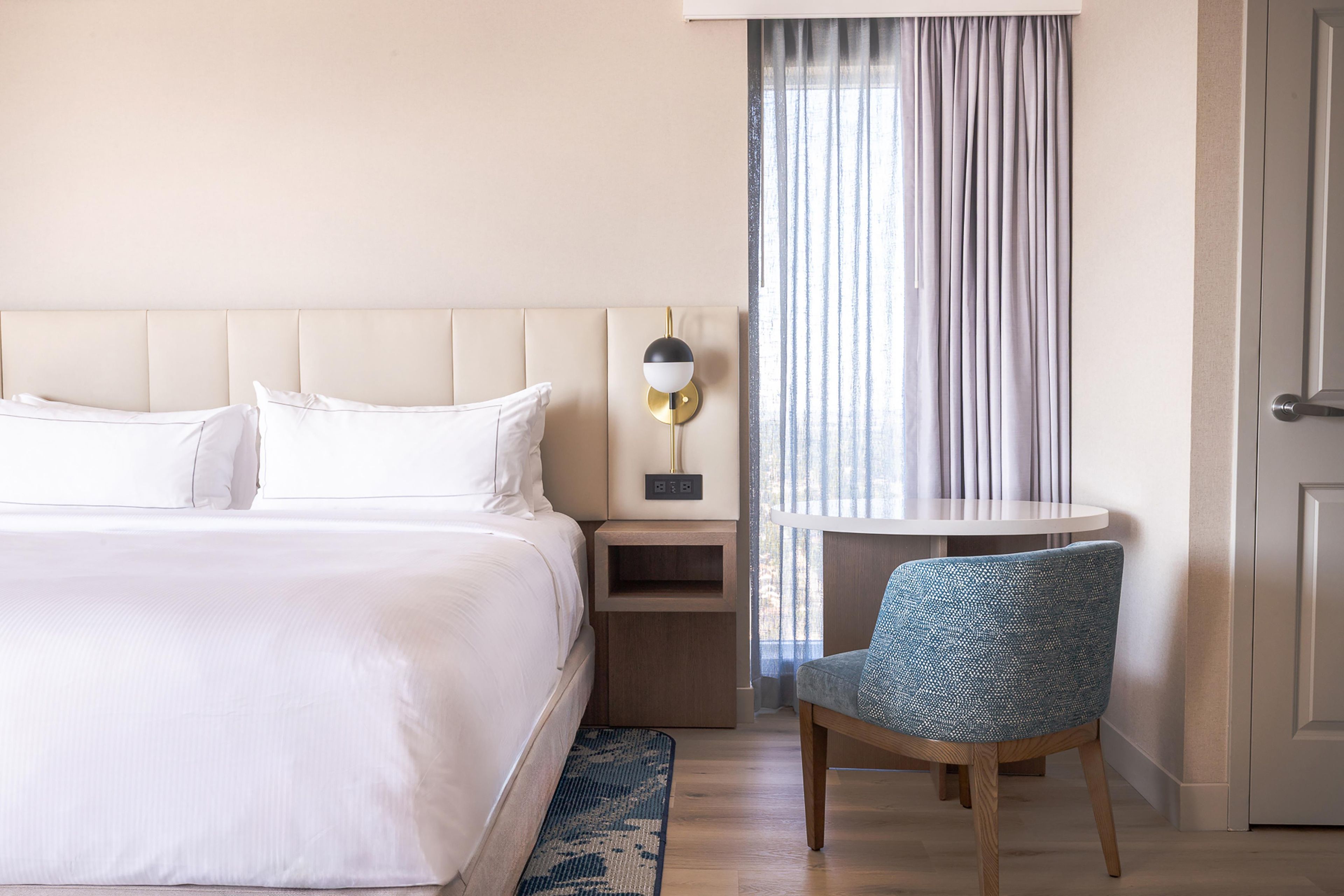 A hotel room with a large bed, a bedside table, a lamp, a chair, and a window with curtains.
