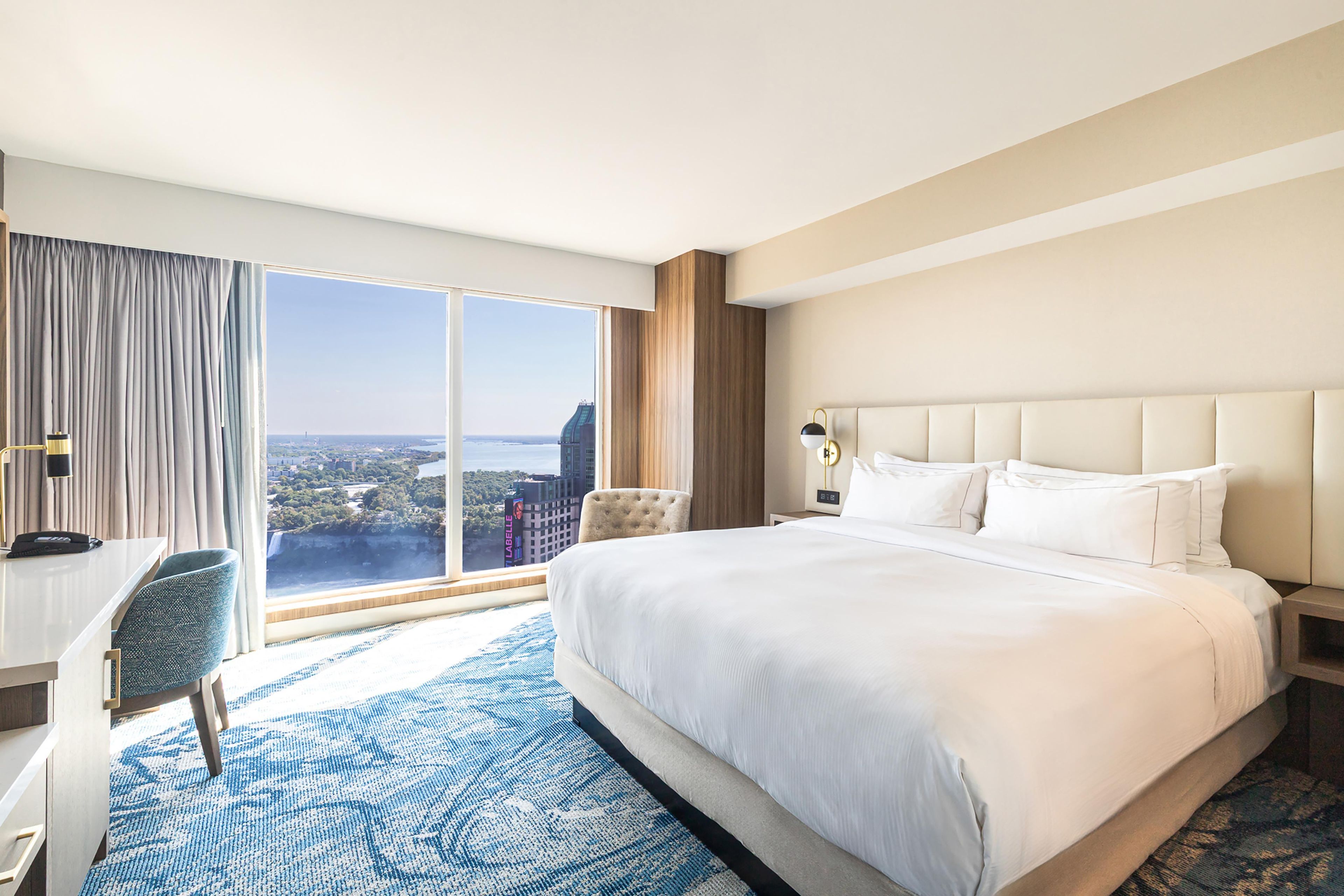 A hotel room with a large bed, a desk, a chair, a view of a cityscape, and a blue patterned rug.