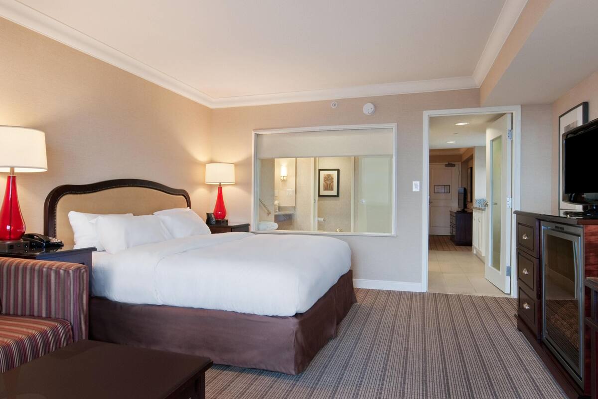 Une chambre d’hôtel comprenant un lit, une table de chevet, une lampe, une télévision, un miroir et une porte donnant accès à une salle de bain.